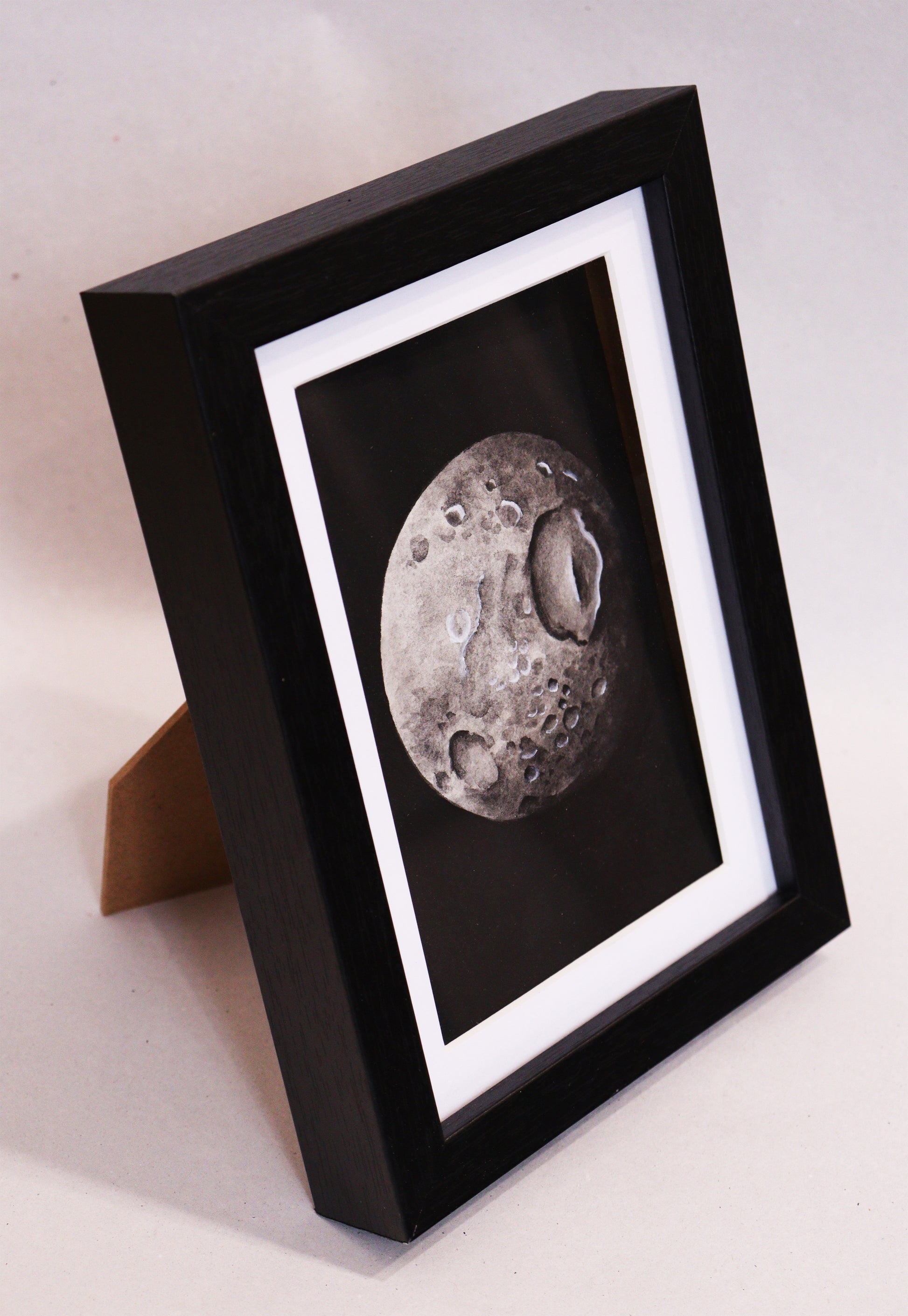 Original Aquarell-Malerei, Gemälde von Lisa Steinbrück, Titel „Mimas“ – Darstellung eines der Monde des Saturns in Aquarell-Stil, schwarz-weiß und Grautöne auf schwarzem Grund, Aquarell auf Papier, gerahmt, schwarzer Rahmen mit weißem Passepartout - Seitenansicht des Rahmens, stehend. Erhältlich bei Galerie Waldbach - What The Artist Made